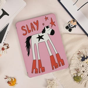 May include: Pink tablet case with a cartoon horse illustration. The horse is white with a black star, mane, and tail, wearing red boots. The word "SLAY" is written in red above the horse, with red stars scattered around.