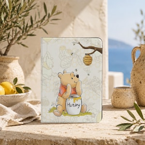 May include: A rectangular tablet case featuring a light-colored background with a Winnie the Pooh illustration. The character is shown sitting and holding a honey pot labeled "HUNNY" with a beehive hanging from a branch above.