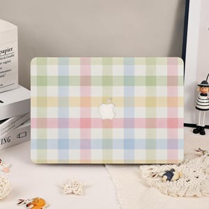 Peut inclure: Une coque d'ordinateur portable avec un motif à carreaux pastel dans des tons doux de rose, jaune, bleu et vert. Le design comprend une découpe centrale qui révèle le logo Apple argenté, créant une esthétique en grille sur la surface.