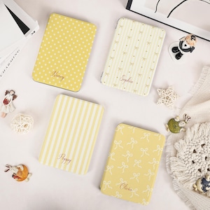 May include: Four rectangular yellow cases with rounded corners. One features white polka dots and the name Nancy. One has white stripes and bows with the name Sophia. One has white stripes and the name Poppy. One has white bows and the name Olivia.
