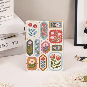 Puede incluir: Una funda rectangular para tableta con fondo color crema decorada con varios motivos florales encerrados en marcos coloridos y ornamentados. El diseño incluye flores rojas, rosas y blancas con hojas verdes dispuestas en una cuadrícula.