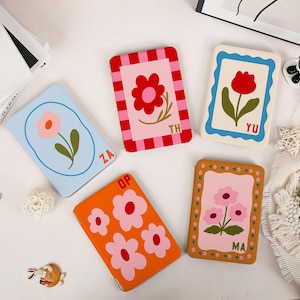 May include: Four rectangular cases featuring floral illustrations. One is light blue with a pink flower and 'ZA', one is red-and-pink striped with a red flower and 'TH', one is cream with a red tulip and 'YU', and one is orange with pink flowers and 'MA'.