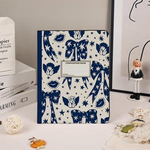 May include: A composition book with a cream-colored background and a navy blue pattern of bows, stars, cherubs, and lips. The cover has a label that reads "COMPOSITION BOOK". The book is standing upright on a white surface.