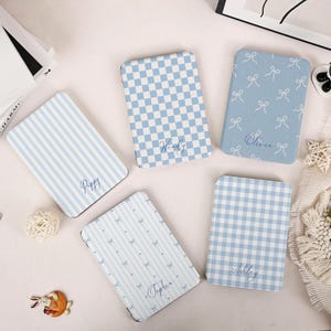 Könnte beinhalten: Vier rechteckige E-Reader-Hüllen in Hellblau und Weiß. Eine zeigt blaue Streifen mit dem Namen Poppy, eine ein blaues Schachbrettmuster mit Wendy, eine ein blaues Schleifenmuster mit Olivia und eine ein blaues Vichy-Muster mit Ashley.