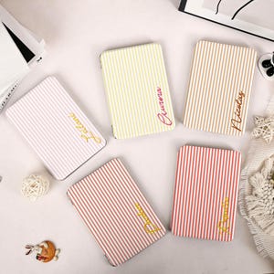 Puede incluir: Cinco estuches rectangulares con rayas verticales en tonos pastel rosa, amarillo, bronceado y coral. Cada estuche está personalizado con un nombre en letra cursiva, incluyendo Leilani, Aurora, Nicolas, Parker y Rosalio, impreso en color dorado u oscuro.