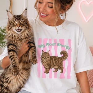 May include: White t-shirt featuring a brown tabby cat graphic and the text "Big Floor Energy" in pink and green. The cat design is set against pink vertical stripes. A person is wearing the shirt and holding a large tabby cat.
