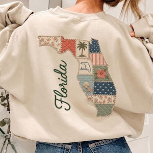 May include: A beige crewneck sweatshirt featuring a large back graphic of the state of Florida. The state shape is filled with a patchwork of patterns including floral, palm tree, hibiscus, and seashell motifs. The word Florida is written in green.