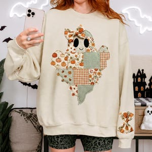 May include: A cream-coloured crewneck sweatshirt featuring a large ghost graphic on the front. The ghost is composed of various patchwork patterns including pumpkins, autumn leaves, and gingham, and wears a bow. A matching bow is on the sleeve.