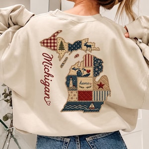 Puede incluir: Una sudadera de cuello redondo color crema con un gran gráfico en la espalda. El diseño muestra el estado de Michigan relleno con un patrón de retazos de varios iconos, incluyendo un faro, un velero y la palabra home en letra cursiva.
