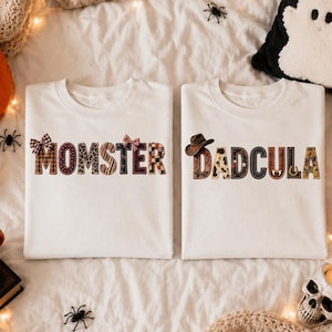 Könnte beinhalten: Zwei weiße T-Shirts mit Rundhalsausschnitt und gemusterten Schriftzügen. Das linke T-Shirt zeigt das Wort MOMSTER mit kleinen Schleifen, das rechte T-Shirt zeigt das Wort DADCULA mit einem Cowboyhut. Beide T-Shirts haben verschiedene Texturen.