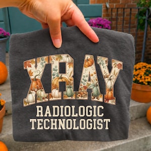 May include: A dark gray t-shirt featuring the text XRAY in large block letters filled with a pattern of autumn leaves and pumpkins. Below, the text RADIOLOGIC TECHNOLOGIST is printed in a cream-colored sans-serif font on the front of the shirt.