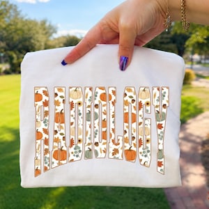 May include: A white t-shirt featuring the word RADIOLOGY in large block letters. The letters are filled with a repeating pattern of orange pumpkins, brown autumn leaves, sunflowers, and green eucalyptus sprigs against a white background.