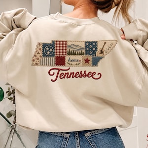 May include: A beige crewneck sweatshirt featuring a large embroidered graphic on the back. The design is shaped like the state of Tennessee, filled with a patchwork pattern of floral, plaid, and mountain motifs, with the word Tennessee in red.