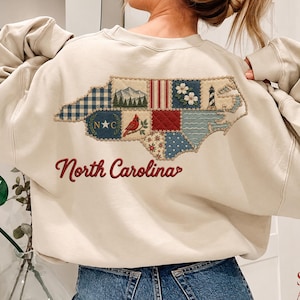 Puede incluir: Una sudadera beige de cuello redondo con un gran diseño bordado en la espalda con la forma de Carolina del Norte. El mapa está compuesto por varios cuadrados de tela estampada que representan montañas, un cardenal, un faro y el texto North Carolina.