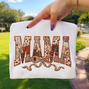 May include: A white t-shirt featuring the word MAMA embroidered in large block letters. The letters are filled with a patchwork pattern of autumn-themed floral, mushroom, and leaf prints in shades of orange, brown, and green, with a bow below.