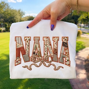 May include: A white t-shirt featuring the word NANA embroidered in large block letters. The letters are filled with a patchwork pattern of autumn-themed fabrics, including mushrooms, ferns, and pinecones, with a decorative bow underneath.