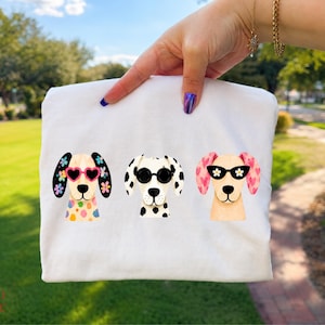 Peut inclure: Un t-shirt blanc avec une impression graphique de trois chiens. Le chien de gauche porte des lunettes en forme de c&oelig;ur et a des oreilles fleuries, celui du milieu a des lunettes rondes noires et des taches, et celui de droite a des lunettes &agrave; fleurs.