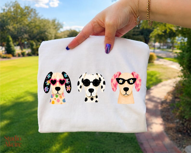 Peut inclure: Un t-shirt blanc avec une impression graphique de trois chiens. Le chien de gauche porte des lunettes en forme de c&oelig;ur et a des oreilles fleuries, celui du milieu a des lunettes rondes noires et des taches, et celui de droite a des lunettes &agrave; fleurs.