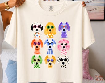 Peut inclure: Un t-shirt à manches courtes de couleur crème avec une impression graphique de neuf chiens disposés en grille de trois par trois. Chaque chien est illustré avec une couleur et un motif différents, portant des lunettes de soleil colorées.