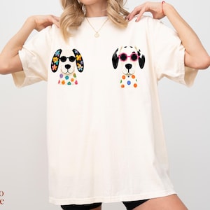 Peut inclure: Un t-shirt &agrave; manches courtes blanc cass&eacute; avec deux illustrations graphiques de chiens dalmatiens. Le chien de gauche porte des lunettes de soleil noires et des fleurs color&eacute;es sur les oreilles et le torse. Celui de droite porte des lunettes roses.