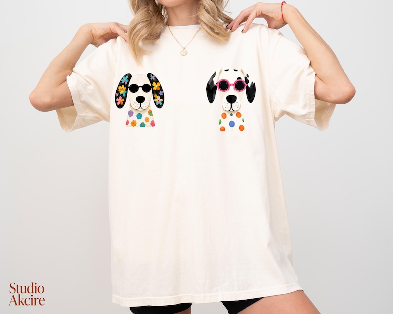 Peut inclure: Un t-shirt &agrave; manches courtes blanc cass&eacute; avec deux illustrations graphiques de chiens dalmatiens. Le chien de gauche porte des lunettes de soleil noires et des fleurs color&eacute;es sur les oreilles et le torse. Celui de droite porte des lunettes roses.