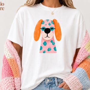 Peut inclure: Un t-shirt blanc &agrave; manches courtes avec une illustration d'un chien rose &agrave; taches bleu sarcelle, oreilles orange et lunettes noires. Le dessin est centr&eacute; sur la poitrine. Le texte Studio Akcire est imprim&eacute; dans le coin sup&eacute;rieur gauche.
