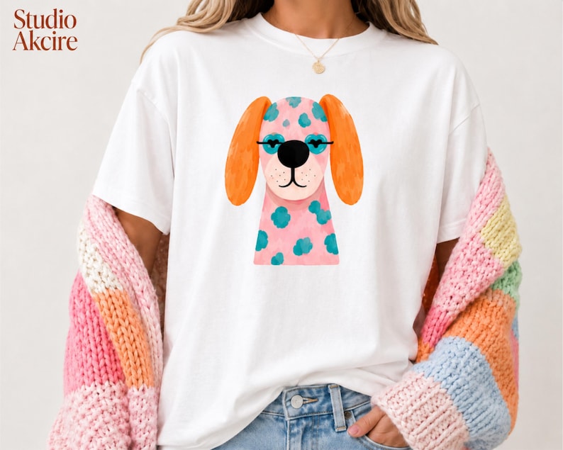 Peut inclure: Un t-shirt blanc &agrave; manches courtes avec une illustration d'un chien rose &agrave; taches bleu sarcelle, oreilles orange et lunettes noires. Le dessin est centr&eacute; sur la poitrine. Le texte Studio Akcire est imprim&eacute; dans le coin sup&eacute;rieur gauche.