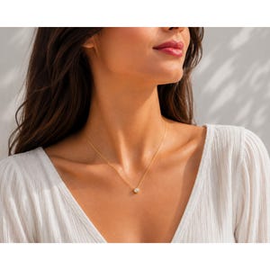 May include: A delicate gold chain necklace featuring a single, small, round, clear gemstone pendant set in a simple gold bezel setting. The necklace rests against the neck, showcasing a minimalist design with a thin, polished gold metal chain.