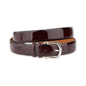 Peut inclure: Une ceinture bordeaux foncé brillante avec une boucle argentée. Enroulée, elle révèle sa finition polie et l'intérieur beige. Un accessoire classique pour compléter diverses tenues.