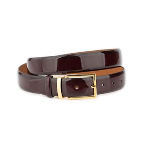 Op de afbeelding: Een glanzende, donker bordeauxrode riem met een goudkleurige rechthoekige gesp. De riem is opgerold en toont zijn gepolijste afwerking en contrasterende bruine binnenkant. De gesp heeft een eenvoudig, elegant ontwerp.