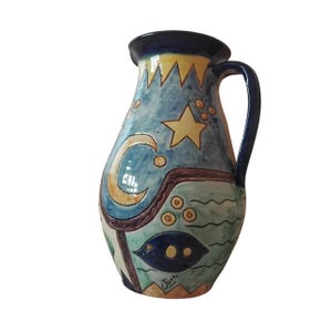 Puede incluir: Jarra de cerámica pintada a mano con un diseño celestial. La jarra presenta un fondo azul con estrellas amarillas, una luna creciente y un pez. El asa y el borde son de color azul oscuro. La firma del artista, "Juan", es visible.