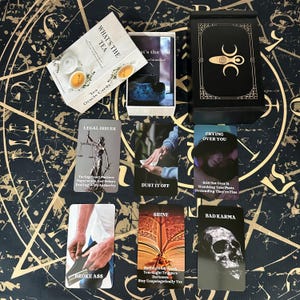 May include: A set of oracle cards with a black and gold box, featuring the text "WHAT'S THE TEA" on the box. Several cards are displayed, with phrases like "LEGAL ISSUES" and "BAD KARMA". The cards are on a patterned surface.