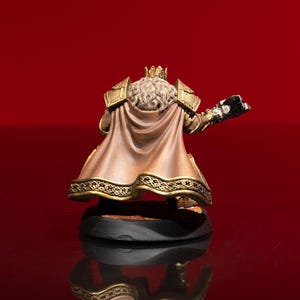 May include: A miniature figurine of a crowned figure wearing gold-colored shoulder armor and a tan cape with a decorative gold-patterned hem. The figure has long, wavy, light-colored hair and is positioned on a circular black base.