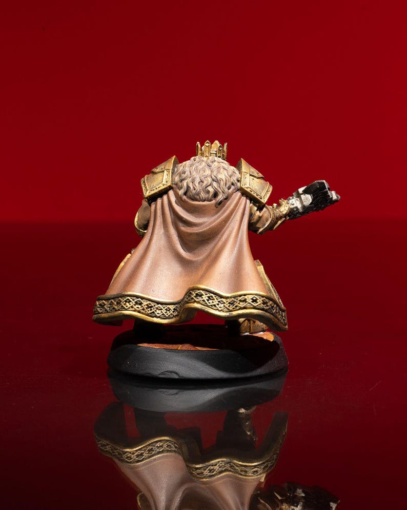 May include: A miniature figurine of a crowned figure wearing gold-colored shoulder armor and a tan cape with a decorative gold-patterned hem. The figure has long, wavy, light-colored hair and is positioned on a circular black base.