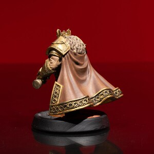 May include: A miniature figurine of a royal figure wearing a gold crown, ornate gold shoulder armor, and a flowing brown cape with intricate gold trim. The figure has long, textured hair and stands on a circular black base, viewed from behind.