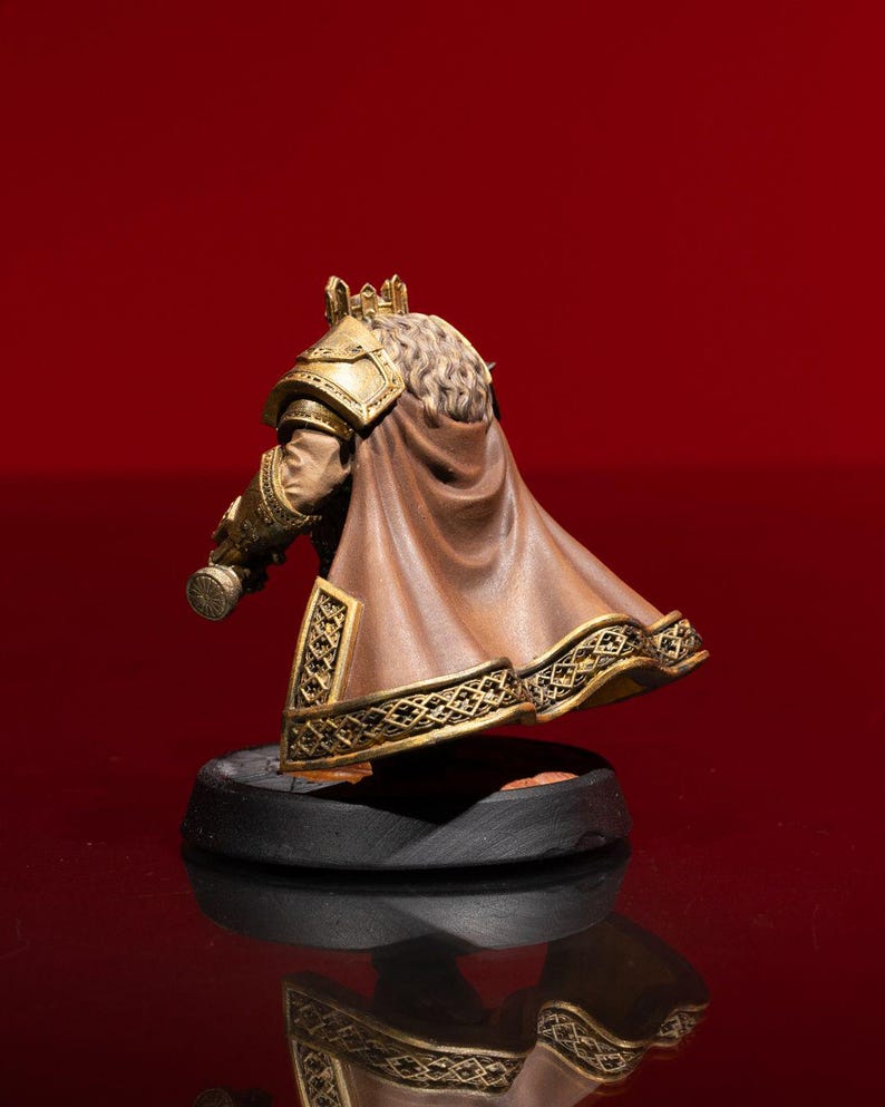 May include: A miniature figurine of a royal figure wearing a gold crown, ornate gold shoulder armor, and a flowing brown cape with intricate gold trim. The figure has long, textured hair and stands on a circular black base, viewed from behind.