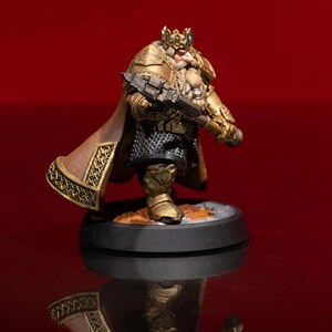 May include: A miniature figurine of a bearded warrior wearing gold-colored plate armor, a crown, and a brown cape. The figure holds a dark, heavy hammer and stands on a circular black base, featuring intricate metallic detailing and chainmail.