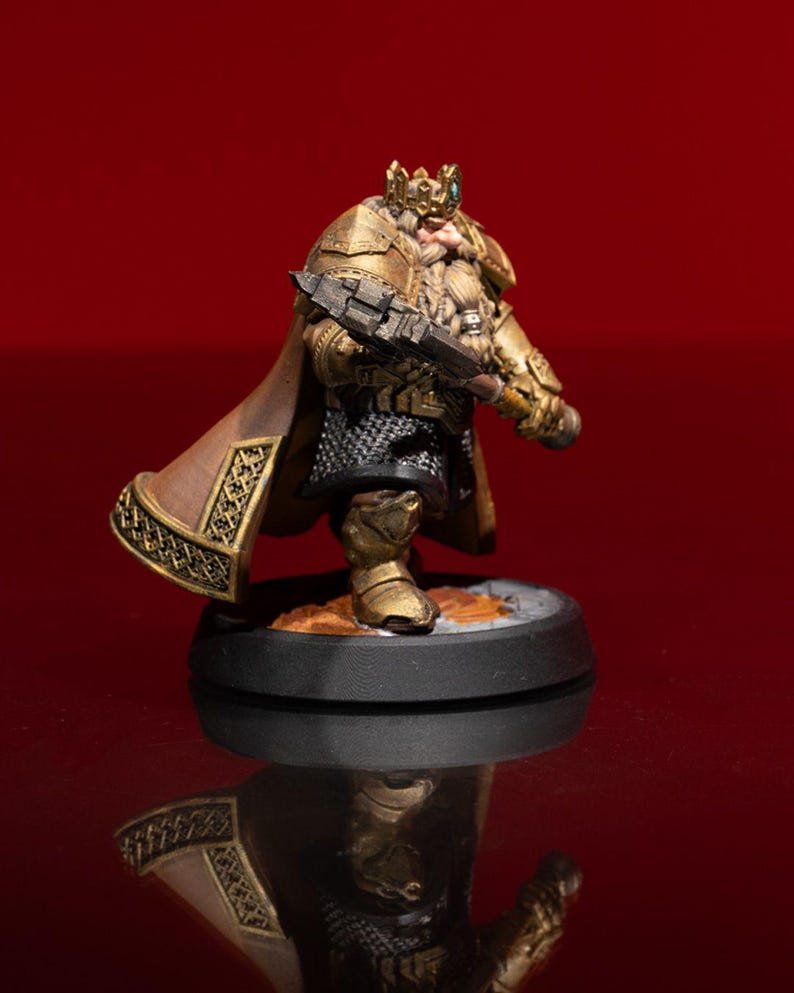 May include: A miniature figurine of a bearded warrior wearing gold-colored plate armor, a crown, and a brown cape. The figure holds a dark, heavy hammer and stands on a circular black base, featuring intricate metallic detailing and chainmail.