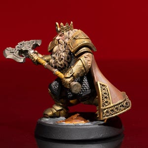 May include: A painted miniature figurine of a bearded dwarf king wearing gold-colored plate armor, a crown, and a brown cloak with intricate gold trim. The figure holds a large silver-toned battle axe and stands on a circular black base.