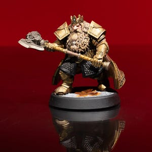 May include: A miniature figurine of a bearded dwarf king wearing gold-colored plate armor and a crown, holding a large battle axe. The figure features intricate chainmail detailing, a long braided beard, and stands on a circular black base.