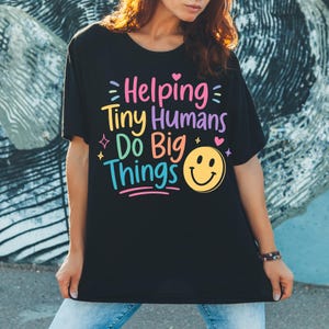 Op de afbeelding: Een zwart t-shirt met de kleurrijke tekst Helping Tiny Humans Do Big Things in roze, geel, paars en blauw, vergezeld door een gele smiley, kleine hartjes en sterpictogrammen gedrukt op de voorzijde van het kledingstuk.