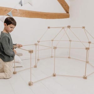 Peut inclure: Un kit de construction en bois clair pour un fort, avec des pièces de connexion. La structure partiellement assemblée est de couleur claire, et un enfant est à genoux à côté. L'arrière-plan est une pièce blanche avec une poutre en bois.