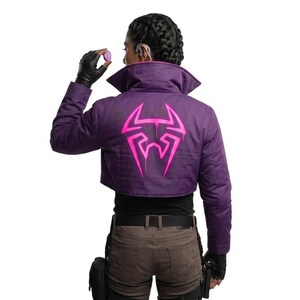 May include: A cropped purple jacket featuring a large, bright pink graphic emblem on the back. The jacket has a high collar, long sleeves, and a structured fit. The pink design resembles a stylized spider or insect motif with sharp, angular lines.