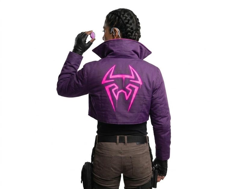May include: A cropped purple jacket featuring a large, bright pink graphic emblem on the back. The jacket has a high collar, long sleeves, and a structured fit. The pink design resembles a stylized spider or insect motif with sharp, angular lines.