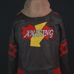 K&ouml;nnte beinhalten: Eine dunkelbraune Lederjacke mit beigefarbener Kapuze, die auf der R&uuml;ckseite eine gro&szlig;e rote und gelbe Grafik mit dem Wort "AMAZING" in wei&szlig;er Schrift zeigt. Die &Auml;rmel haben rote sternf&ouml;rmige Akzente und die Jacke hat einen roten gerippten Saum und B&uuml;ndchen.