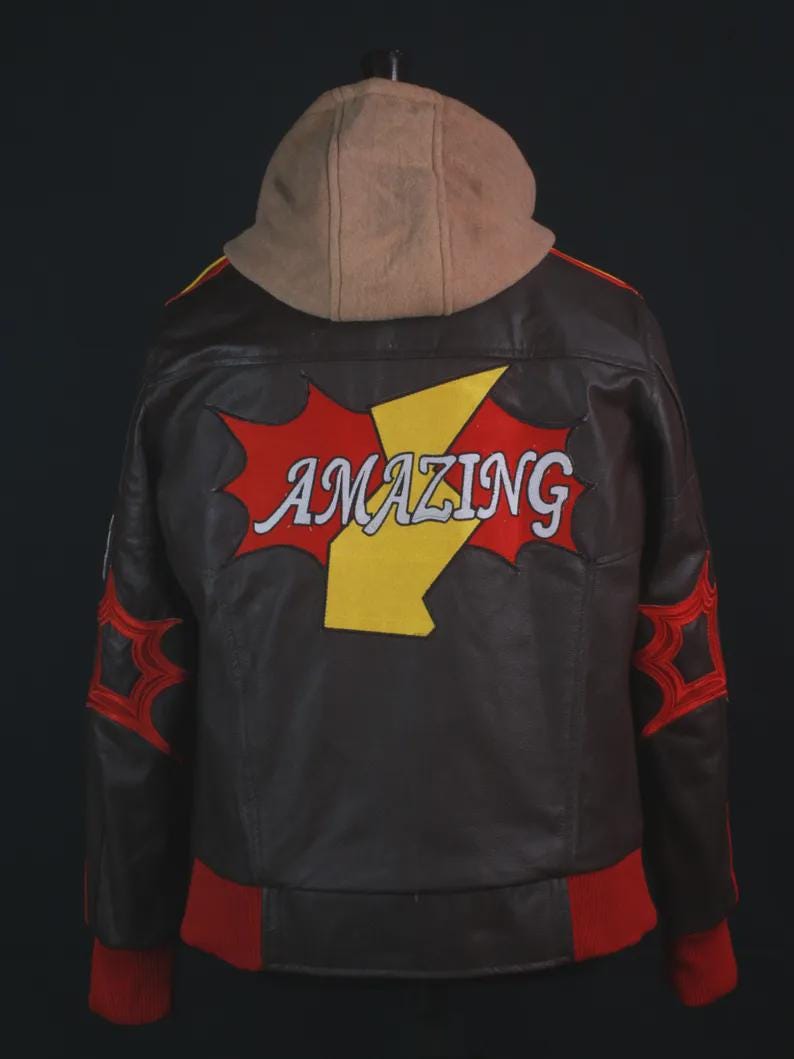 K&ouml;nnte beinhalten: Eine dunkelbraune Lederjacke mit beigefarbener Kapuze, die auf der R&uuml;ckseite eine gro&szlig;e rote und gelbe Grafik mit dem Wort "AMAZING" in wei&szlig;er Schrift zeigt. Die &Auml;rmel haben rote sternf&ouml;rmige Akzente und die Jacke hat einen roten gerippten Saum und B&uuml;ndchen.