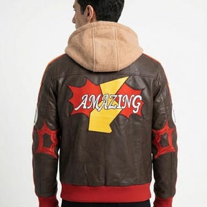 K&ouml;nnte beinhalten: Eine braune Lederjacke mit beigefarbener Kapuze, rotem Rippsaum und roten Rippb&uuml;ndchen. Auf der R&uuml;ckseite befindet sich ein gro&szlig;es rot-gelbes Grafik-Patch mit dem Wort AMAZING in wei&szlig;er Schrift sowie rote sternf&ouml;rmige Aufn&auml;her an den &Auml;rmeln.