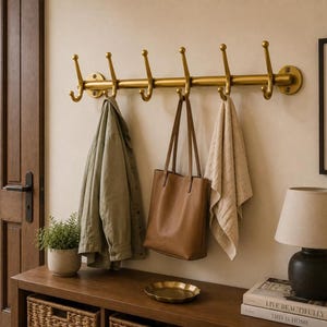 May include: A wall-mounted brass coat rack featuring six double hooks arranged in a horizontal line. The rack is secured to the wall with circular plates at each end. A light green jacket, a brown leather tote bag, and a beige towel hang from it.