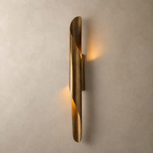 May include: A gold-colored, wall-mounted sconce with a curved, tubular design. The sconce has a warm, inviting glow emanating from within its curved form. The fixture is made of metal and is mounted on a neutral-colored wall.