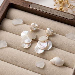May include: A pair of dangle earrings featuring iridescent white shell discs suspended from gold wire-wrapped square shell beads. The earrings are displayed in a wooden box with a metal plaque reading Lunar Winds Artisanal Jewelry - White Shell Cascades.